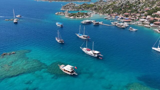 Aerial forward tracking shot over the turquoise waters of Kekova bay and Kalekoy village, showing anchored boats, sunken ruins, and the ancient castle. (Turkuaz Kekova koyu ve Kalek&ouml;y k&ouml;y&uuml; &uuml;zerinde il