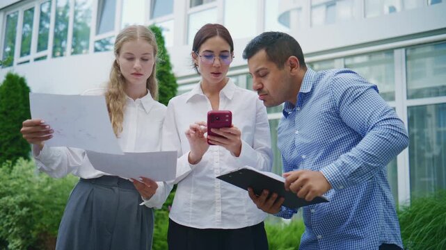 Business meeting takes place outdoors with three people discussing documents and using a phone for information