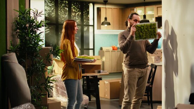 Man and woman unpack wrapped household items from cardboard boxes while redecorating a new house, hanging decorations and arranging belongings as they settle into their first home.