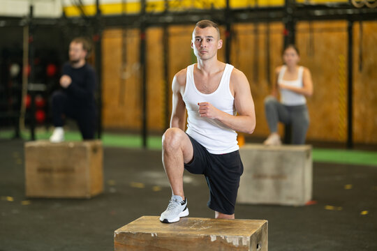 Focused young adult man performing step-ups on plyo box to enhance lower body strength in functional training session with group at gym