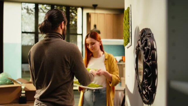 Couple arranges decorations and paintings on the wall in a new house, unpacking belongings from cardboard boxes during relocation day as homeowners create a cozy and personal forever home.