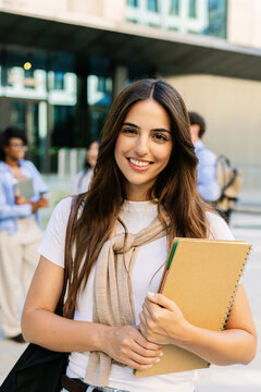 Portrait of a happy young caucasian female business school student smiling and holding a notebook outdoors, MBA classmates blurred in background.