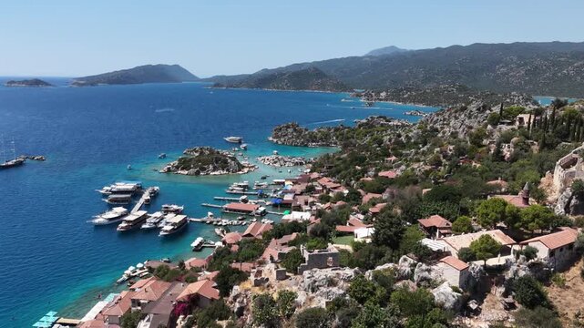 Cinematic aerial parallax shot orbiting the historic coastal village of Kalekoy (Simena) and its harbor with anchored boats.