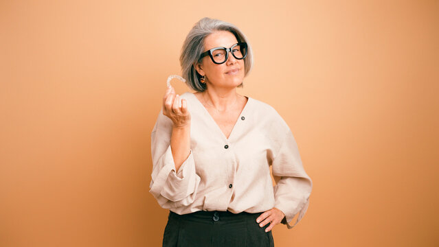 Adult woman with grey hair and glasses presenting a transparent invisible brace for teeth straightening, promoting modern dentistry