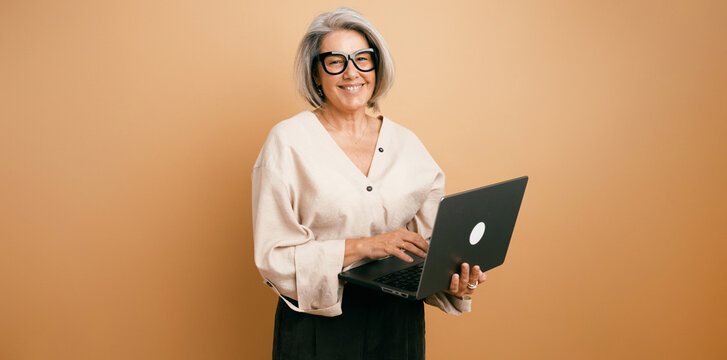 Senior woman wearing glasses standing against a plain background, actively using a laptop and smiling