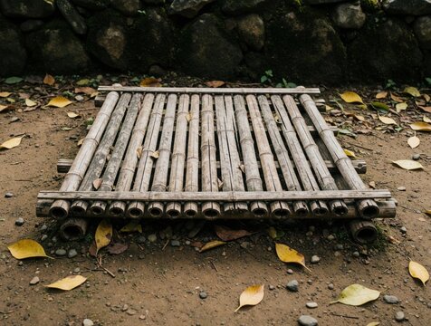 Weathered bamboo platform rests on dirt ground near mossy stone wall. Fallen yellow leaves scatter around rustic Asian garden decor. Simple, aged wood structure lies outdoors.