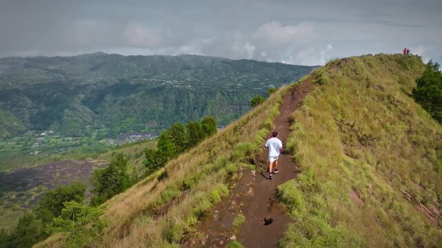 Person hiking on a narrow mountain ridge, enjoying the panoramic views of the volcanic landscape, green valleys, and distant villages under a cloudy sky in Bali. Aerial view drone panorama