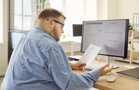Overweight man checks electricity bill on computer. At a home desk he compares screen totals with documents, plans a budget, and schedules online payment. Household finance and budget concept.