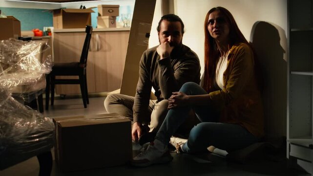 Couple sitting on the floor during a break from relocation, surrounded by cardboard boxes in an empty new house while discussing furniture layout and enjoying the satisfaction of moving in together.