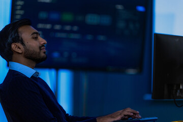 Fototapeta premium A male data analyst in a dark navy suit jacket and light blue shirt watches a chart update while deciding whether to escalate in a blue lit monitoring room in a modern technology company