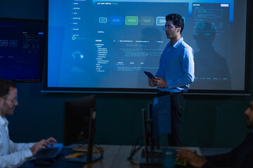 Fototapeta premium A young male analyst with dark curly hair and clear framed glasses in a light blue shirt raises an open palm to emphasise a point about network security in a blue lit monitoring room