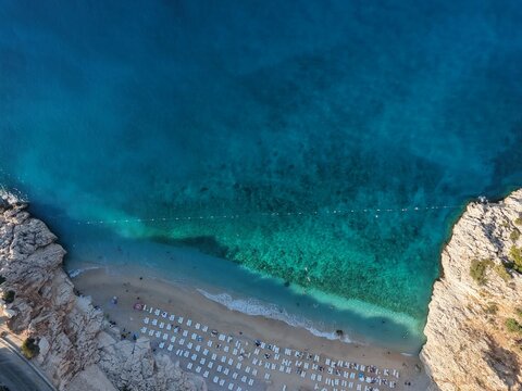 Aerial top-down view of a stunning sandy beach nestled between steep rocky cliffs with crystal clear turquoise water and white sun loungers.