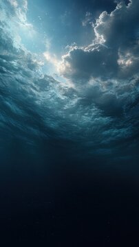 Mysterious view from beneath the ocean surface revealing dramatic cloudy sky and sunlight penetrating deep blue water creating a serene underwater atmosphere