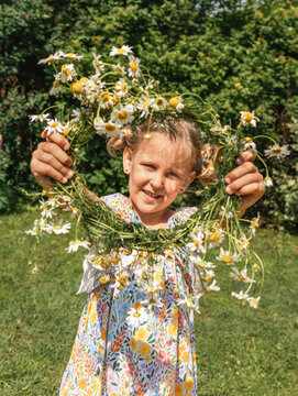 Little girl smiling and holding a handmade chamomile wreath in sunny summer garden, embracing childhood happiness. Real childhood, outdoor play, curiosity and connection with nature