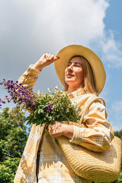 Woman in straw hat with basket of wildflowers including lupines and daisy enjoys sunny day outdoors. Rural lifestyle, flower picking, slow living and connection with nature