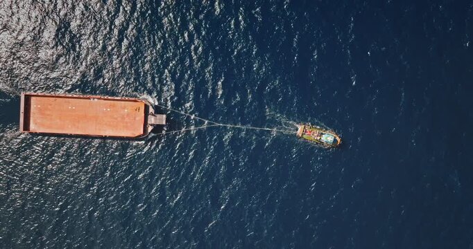 Tugboat pulling large cargo ship, navigating blue ocean. Water transportation concept. Aerial top down drone zoom out shot