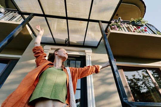 Happy young woman standing on a terrace raises her arms with joy near a modern house. She wears an orange shirt and green top. Concept of freedom, new home and happy lifestyle.