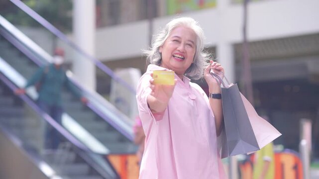 Portrait of Happy Asian active senior woman holding credit card and shopping bag at shopping mall. Elderly retired woman enjoy urban lifestyle shopping with using contactless payment, online banking.