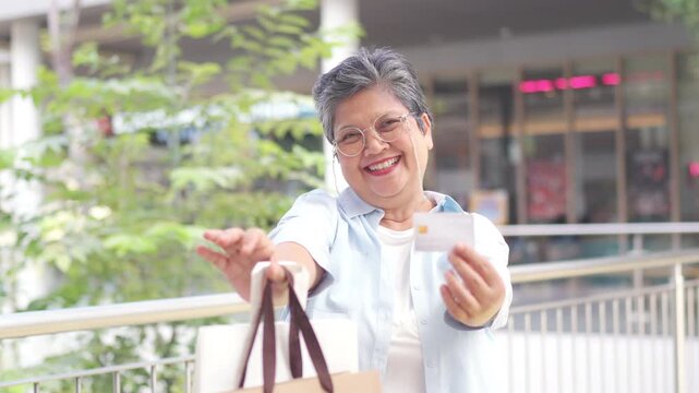 Portrait of Happy Asian active senior woman holding credit card and shopping bag at shopping mall. Elderly retired woman enjoy urban lifestyle shopping with using contactless payment, online banking.
