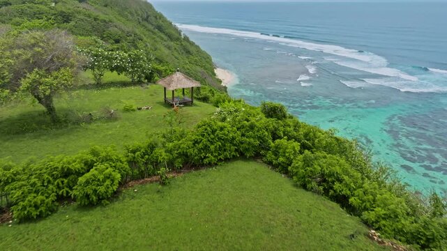 Gazebo on a green clifftop overlooking the expansive turquoise ocean, featuring coral reefs, gentle waves and a serene sandy beach along the tropical coastline of Amed, Bali. Tropical nature landscape