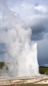 Famous Old Faithful cone geyser erupting on a sunny day with dark clouds in background, summer tourism at Yellowstone National Park viewing natural geothermal features

