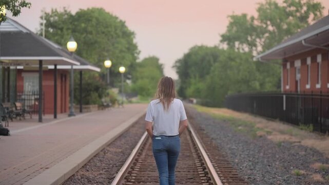 A young woman with long hair walks away from camera down the center of train tracks at a small town depot, lit lamps and red brick buildings on either side under a pastel sunrise sky.