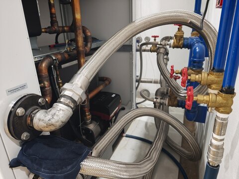 A close-up shot of an intricate industrial plumbing and piping assembly, featuring brass gate valves with red handwheels, blue metal pipes, and flexible stainless steel braided hoses.