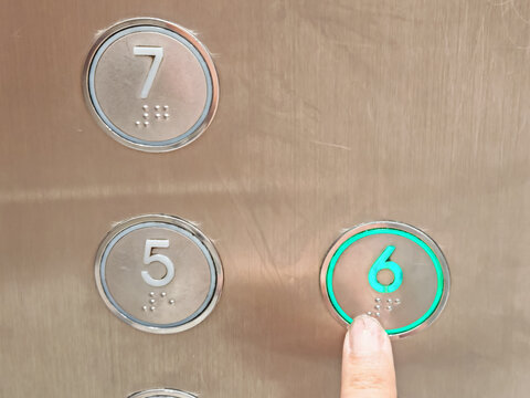 a person's finger pressing the glowing green elevator button for the sixth floor featuring brushed metal paneling and braille accessibility characters.