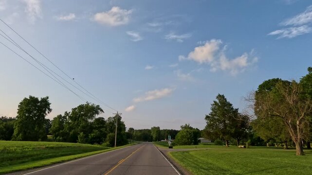 Point of view shot traveling down a winding rural road in Midwest America. Captures classic country living with farmhouses, roadside greenery, an oncoming car, and trucks displayed outdoors.