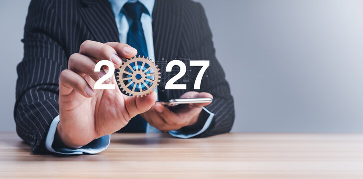 Businessman holding wooden gear and smartphone with 2027 new year numbers. Operations strategy, automation, process optimization, enterprise technology, efficiency, and digital transformation concept.