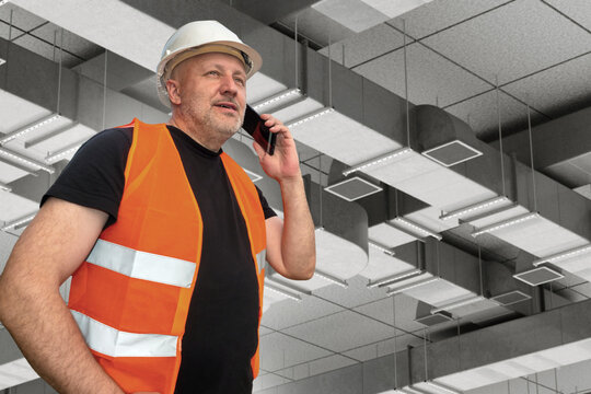 HVAC engineering. Building services. Engineer talks on a mobile phone while inspecting ventilation and air conditioning ductwork during commercial building installation.