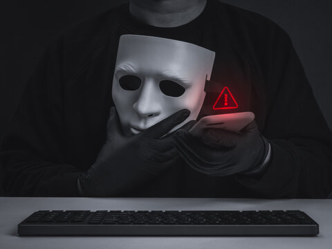 Dark conceptual image of masked cybercriminal scammer holding smartphone beside red warning icon and keyboard. Digital identity theft, phishing, fraud, mobile security, and account takeover concept.