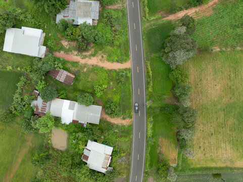 Drone view of paved country road between farmland and village homes during wet growing season, showing rural infrastructure that connects farming communities, agriculture and local food production.