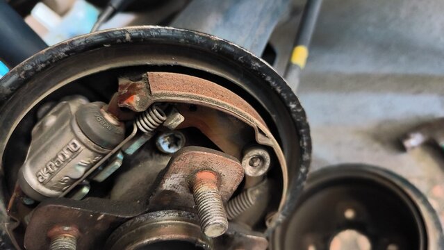 Worn brake assembly sits exposed in garage setting. Metal parts show signs of use and age. Rust, grease, and wear mark all surfaces. Mechanic's tools nearby imply recent work