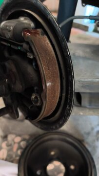 Worn brake assembly sits exposed in garage setting. Metal parts show signs of use and age. Rust, grease, and wear mark all surfaces. Mechanic's tools nearby imply recent work