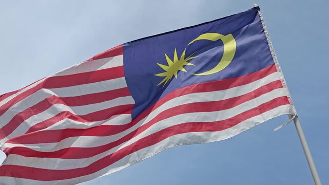 The national flag of Malaysia, Jalur Gemilang, flutters proudly against a clear blue sky, held by Homo sapiens