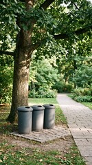 Obraz premium Three grey garbage bins stand neatly on a paved pad beside a walking path under a large leafy tree in a peaceful public park