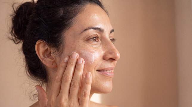 A Hispanic woman applies moisturizer in soft warm light for a skincare article banner.