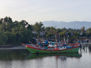 Fototapeta premium Fishing Boat Moored on River with Lush Green Trees and Distant Mountains.