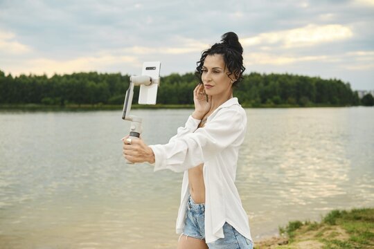 Woman stands by lake holding smartphone. She smiles as she captures photo near water. Trees are visible in the background under cloudy sky