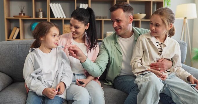 Family parents mother father and daughters bonding together. They sit on a sofa at home, smiling, chatting, and enjoying quality time. Great for lifestyle campaigns. Theme of love family unity.