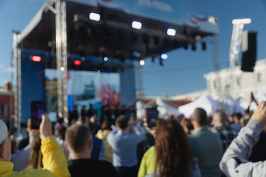 Back view of an audience watching live concert in blur or public event on outdoor stage setup on sunny day, with people holding up smartphones to capture show