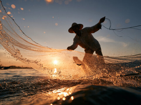 Fisherman casting a large fishing net at golden sunset over the water. Silhouette with sun flare and water splashes in traditional net fishing.