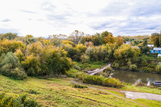 Rural landscape with a temporary bridge over a narrow river surrounded by autumn forest and green field, cloudy day scene.