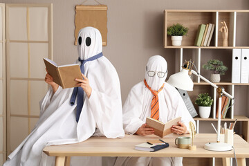 Two people dressed as ghosts with ties reading books at an office desk, halloween and business