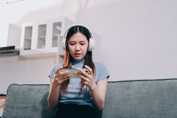 Beautiful young asian woman sitting on sofa and holding joystick playing online video game in living room at house © ARMMY PICCA