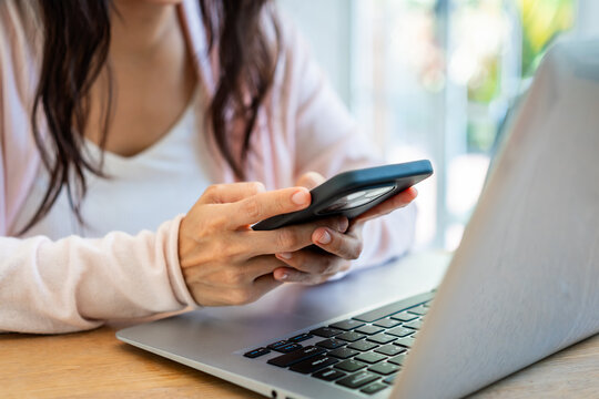 Asian female freelancer holding a mobile phone, messaging or checking notifications in front of a laptop