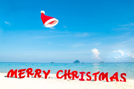 Santa hat floating above 'Merry Christmas' on a sunny beach.