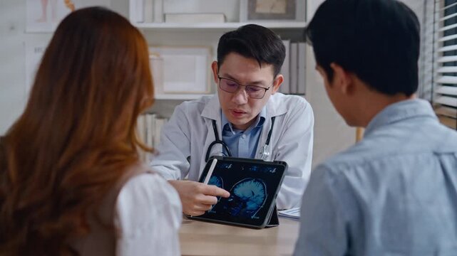 Professional Asian male neurologist doctor explaining brain MRI scan results on digital tablet to a Couple - relationship in modern clinic. Concept of healthcare, diagnosis, and medical technology.