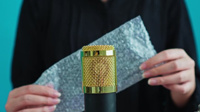 Hands popping bubble wrap behind gold condenser microphone on cyan background